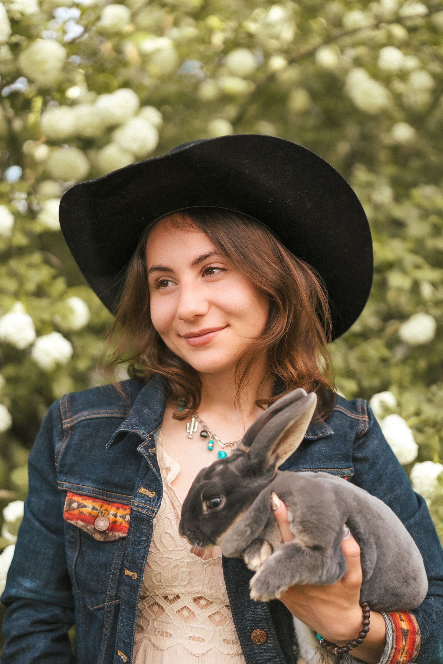 A person in denim jacket and black hat holds two adorable grey rabbits.