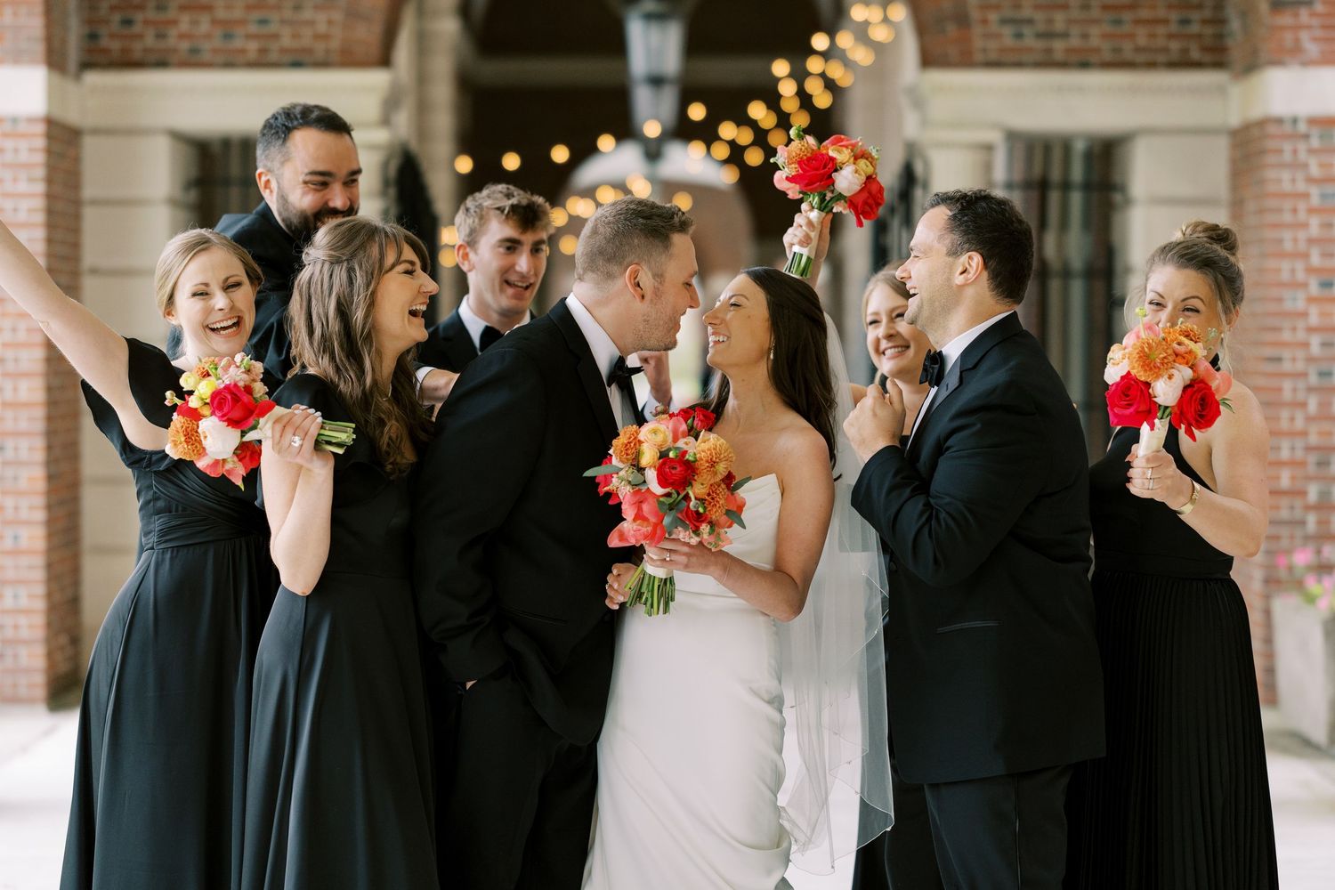 Wedding party in black and navy attire laughs together while holding coral bouquets at brick venue with twinkling lights at Hall of Springs.