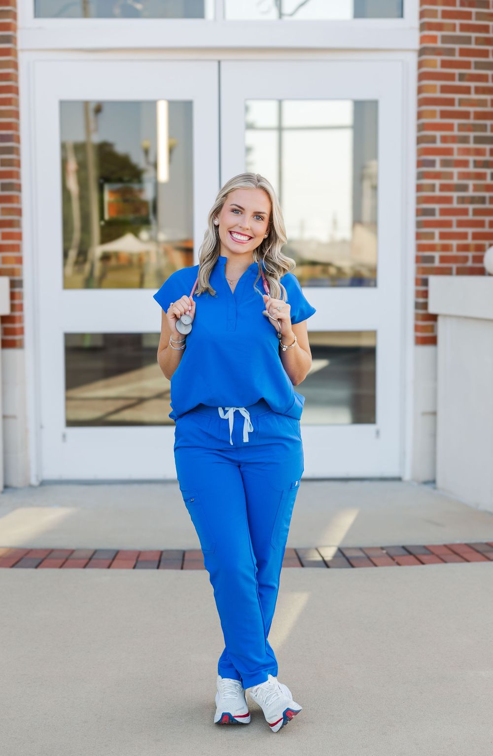 Healthcare professional in bright royal blue scrubs stands smiling outside a brick building entrance with glass doors.