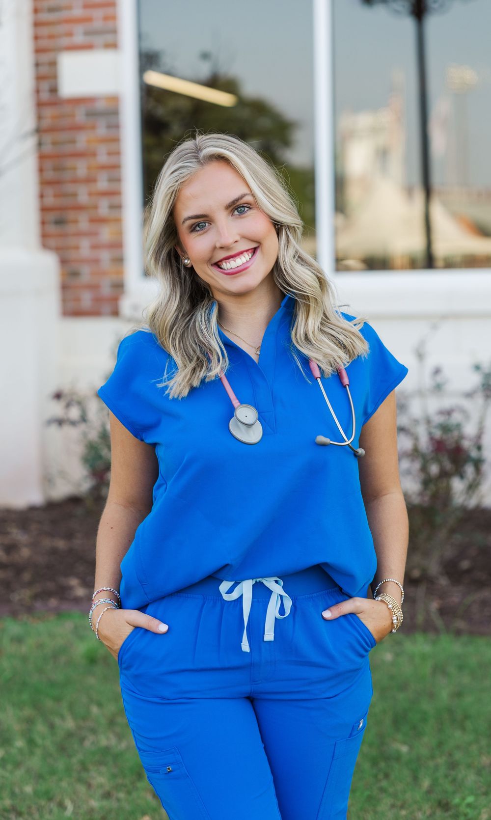 Healthcare worker in bright blue medical scrubs with stethoscope smiling outdoors by brick building.