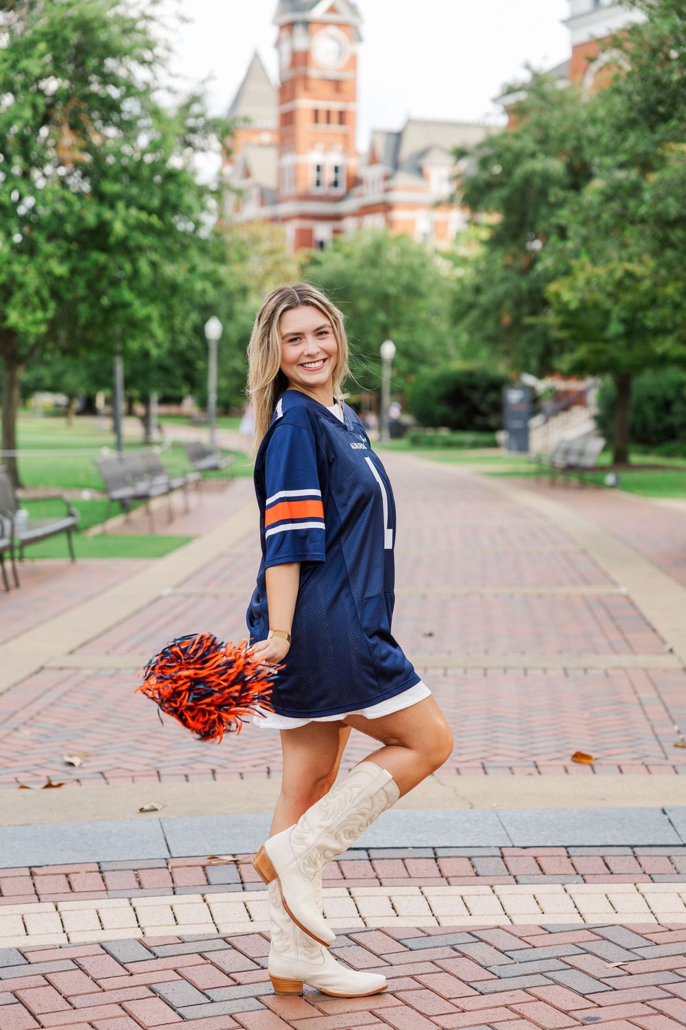 Cheerleader in navy jersey and boots poses with orange pom poms on brick pathway with historic building in background.