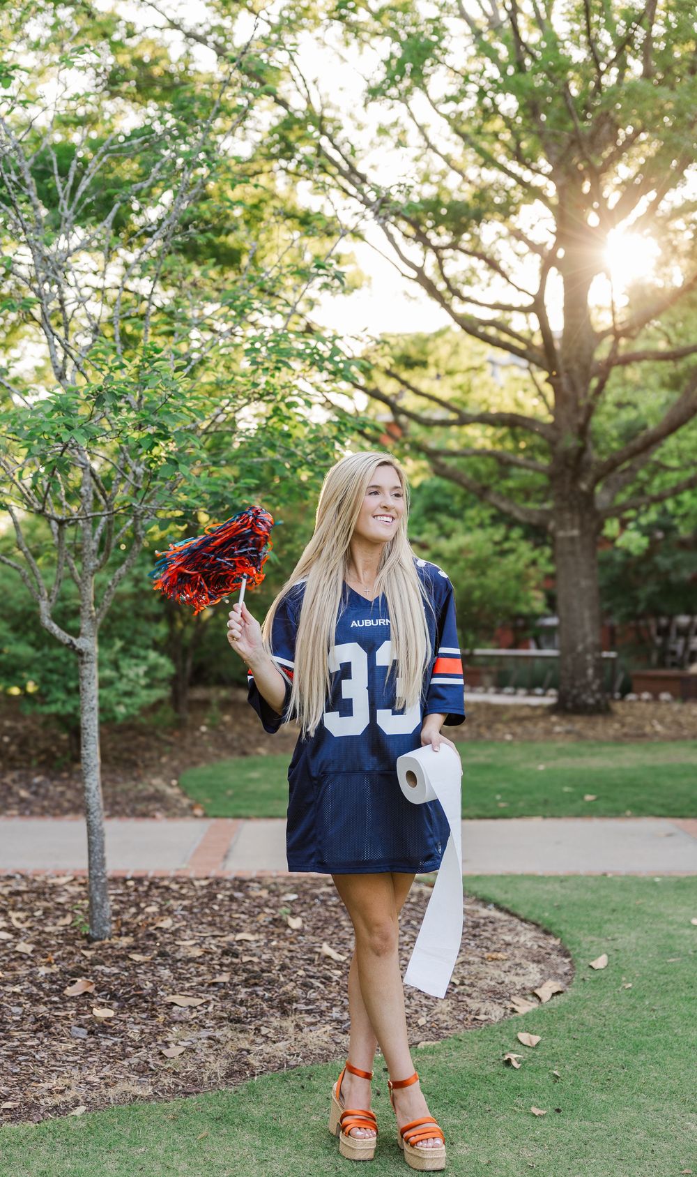 A sports fan stands outdoors holding red flowers and wearing a blue football jersey number 33 and orange sandals at sunset.