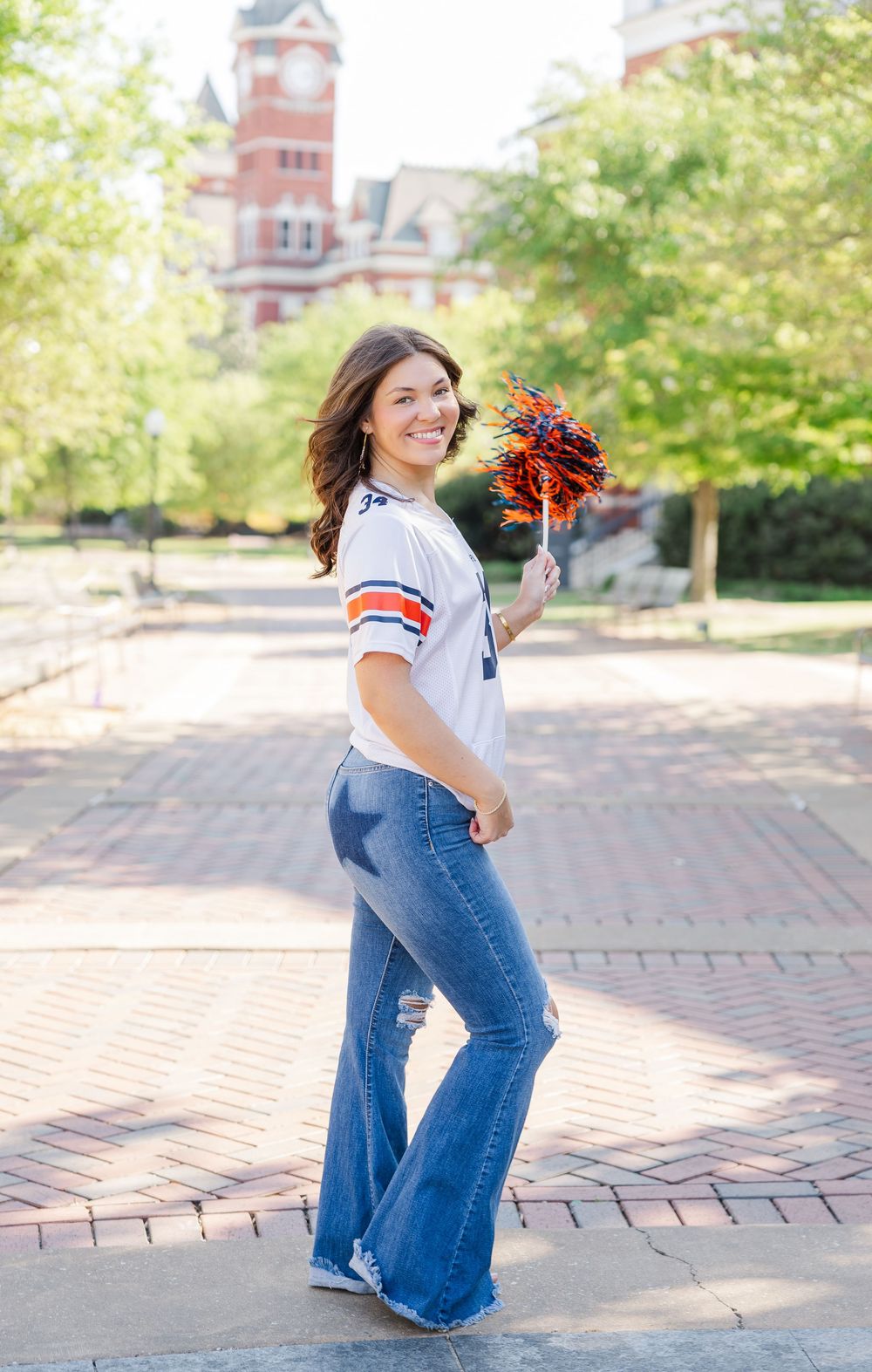 Cheerful person in bell-bottom jeans and white striped top holding orange pom poms on brick walkway near historic building.