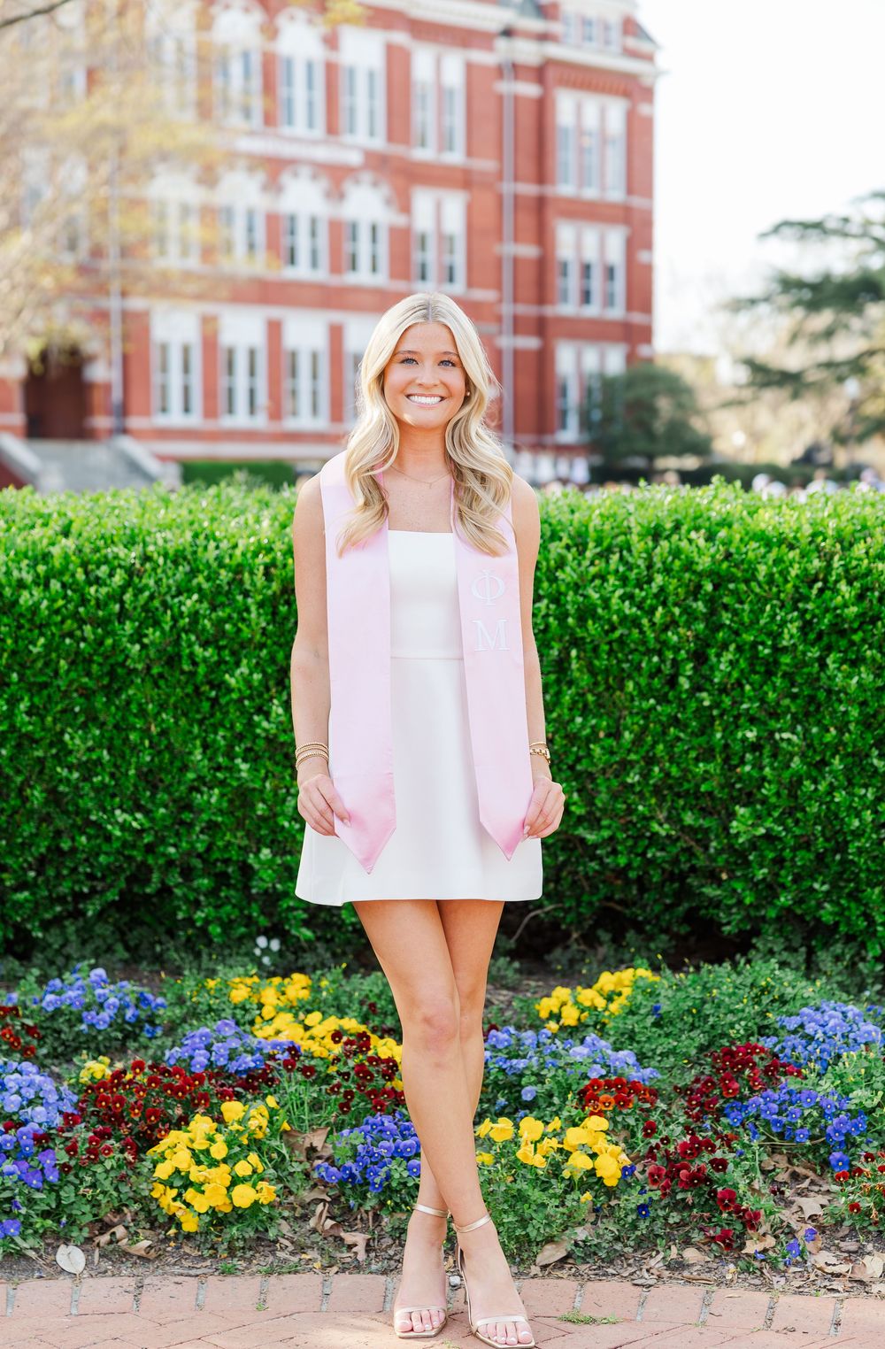 A pink cardigan over a white dress is styled with floral landscaping and a red brick building in the background.