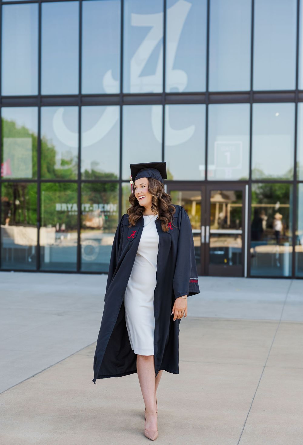 Graduate wearing black cap and gown with white dress and beige heels poses in front of modern glass building.