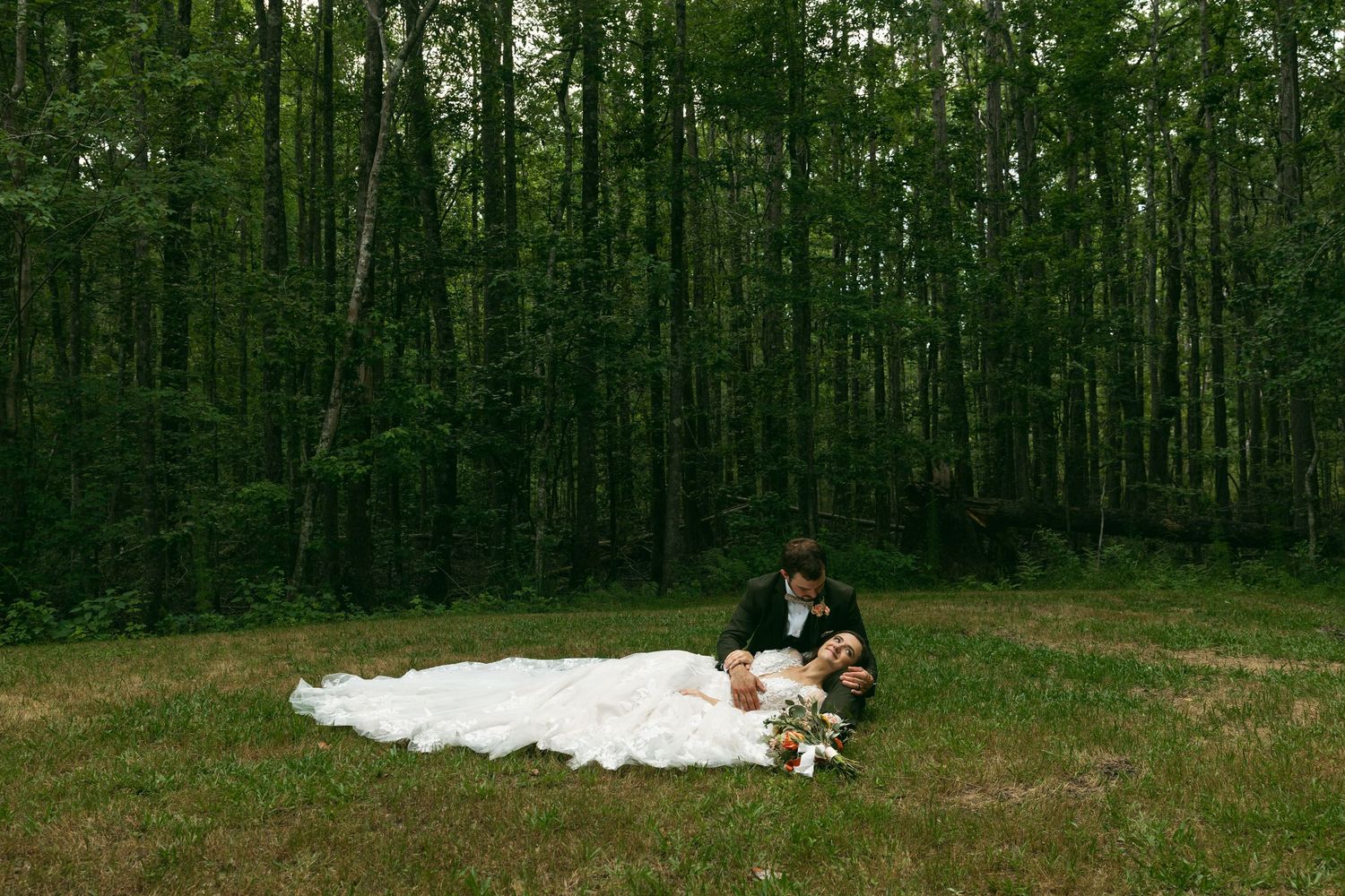 A long white dress spreads across a green grass clearing in front of a deep forest backdrop.