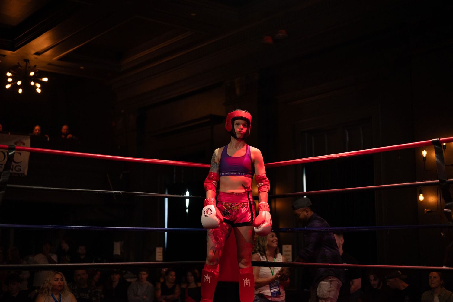 A boxer in red gear stands in a dimly lit boxing ring with ropes illuminated by overhead lights.