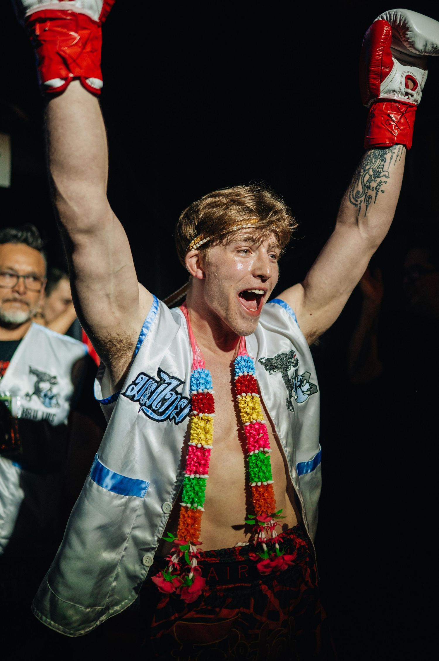 A boxer celebrates victory in the ring wearing a white satin jacket, red boxing gloves and colorful scarf with arms raised.