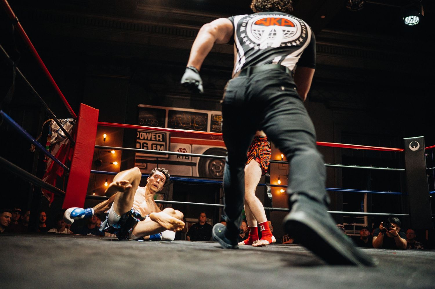 Professional wrestling match in a dimly lit ring with a referee watching as two wrestlers compete on the canvas.