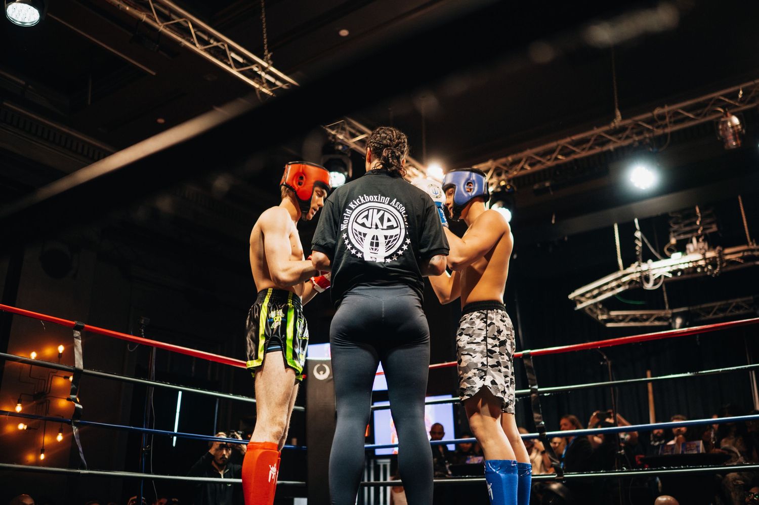 Three kickboxers stand in a boxing ring during a match with stage lighting and equipment visible overhead.