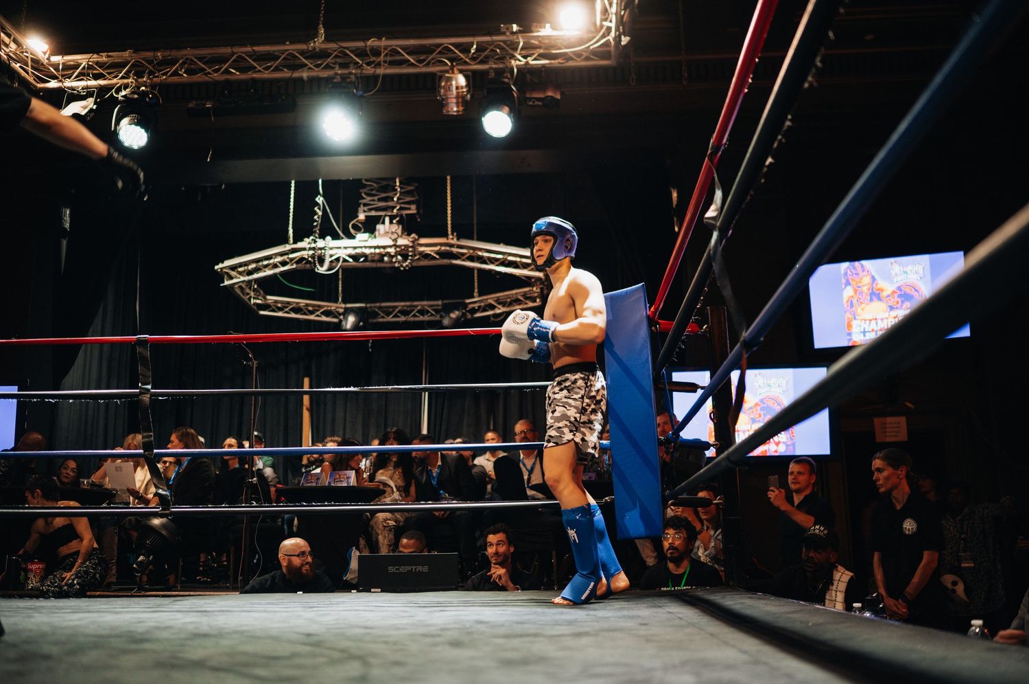 A wrestler in blue tights performs in a wrestling ring under bright spotlights as a crowd watches from the darkness.