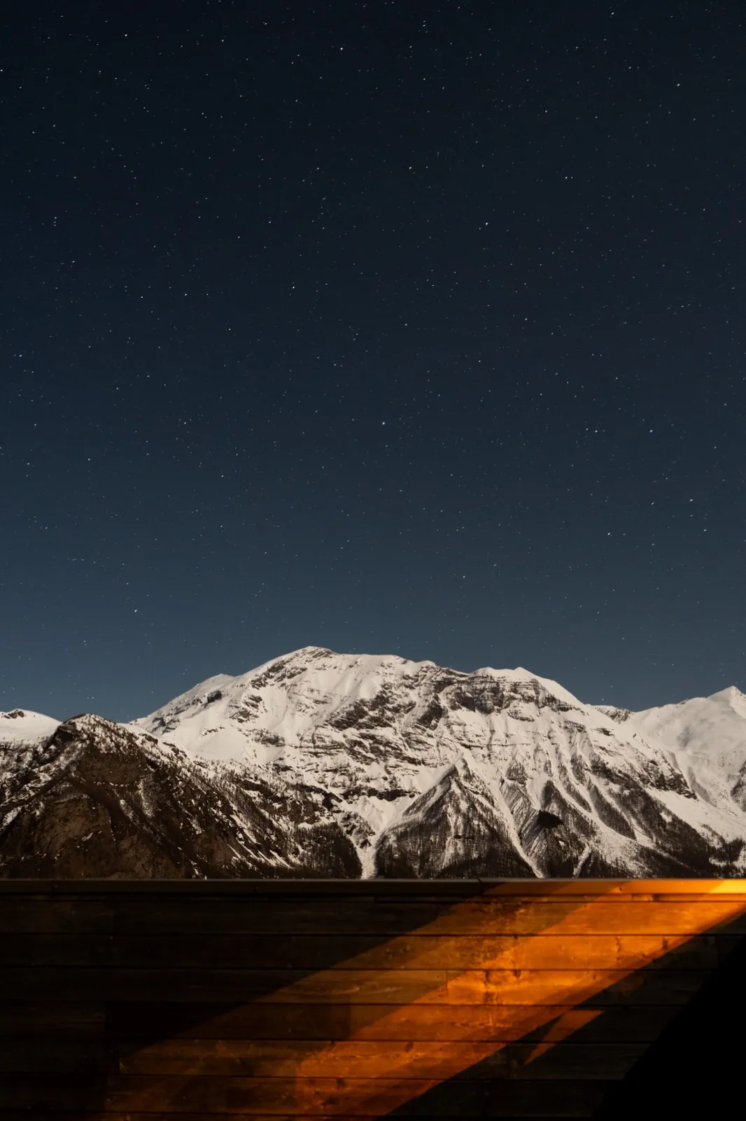 Chaîne de montagnes enneigées sous un ciel nocturne étoilé, avec une lumière orangée éclairant la vallée au premier plan.
