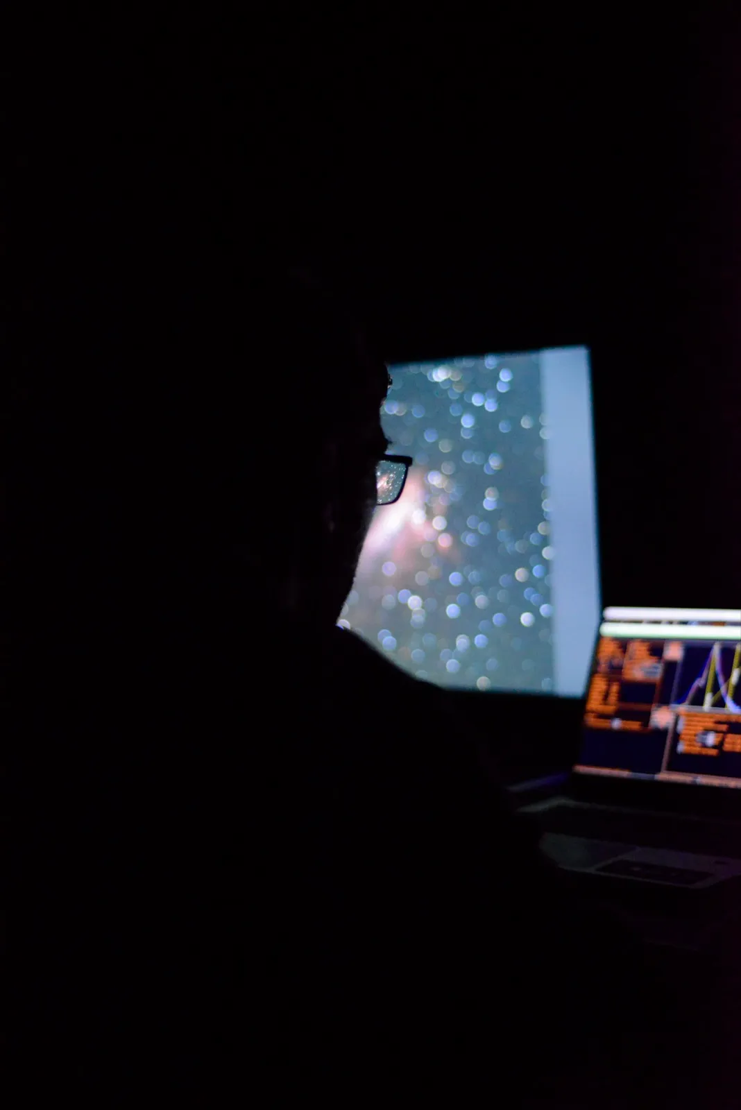 Silhouette dans l’obscurité, face à un écran montrant une nébuleuse ou un amas d’étoiles, probablement en train de contrôler un télescope.