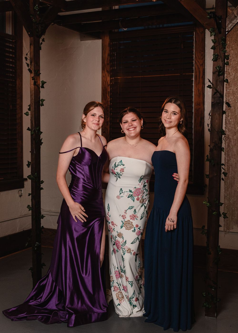 Three friends pose during a formal event wearing elegant dresses in purple, floral, and navy blue.