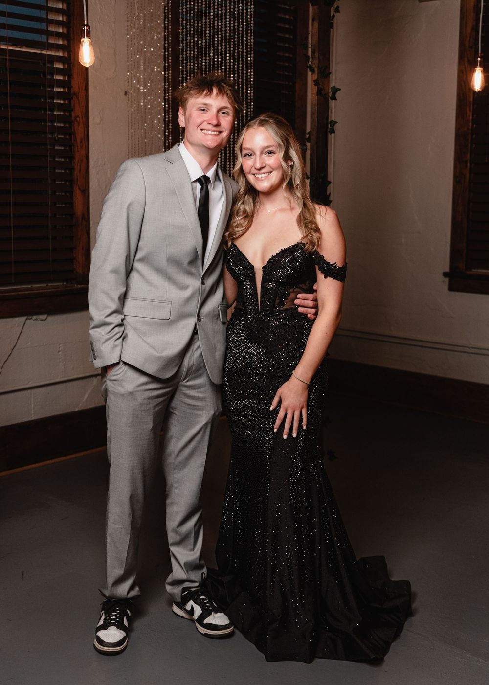 A couple poses together at a formal event, with one person in a grey suit and the other in a sparkly black gown.