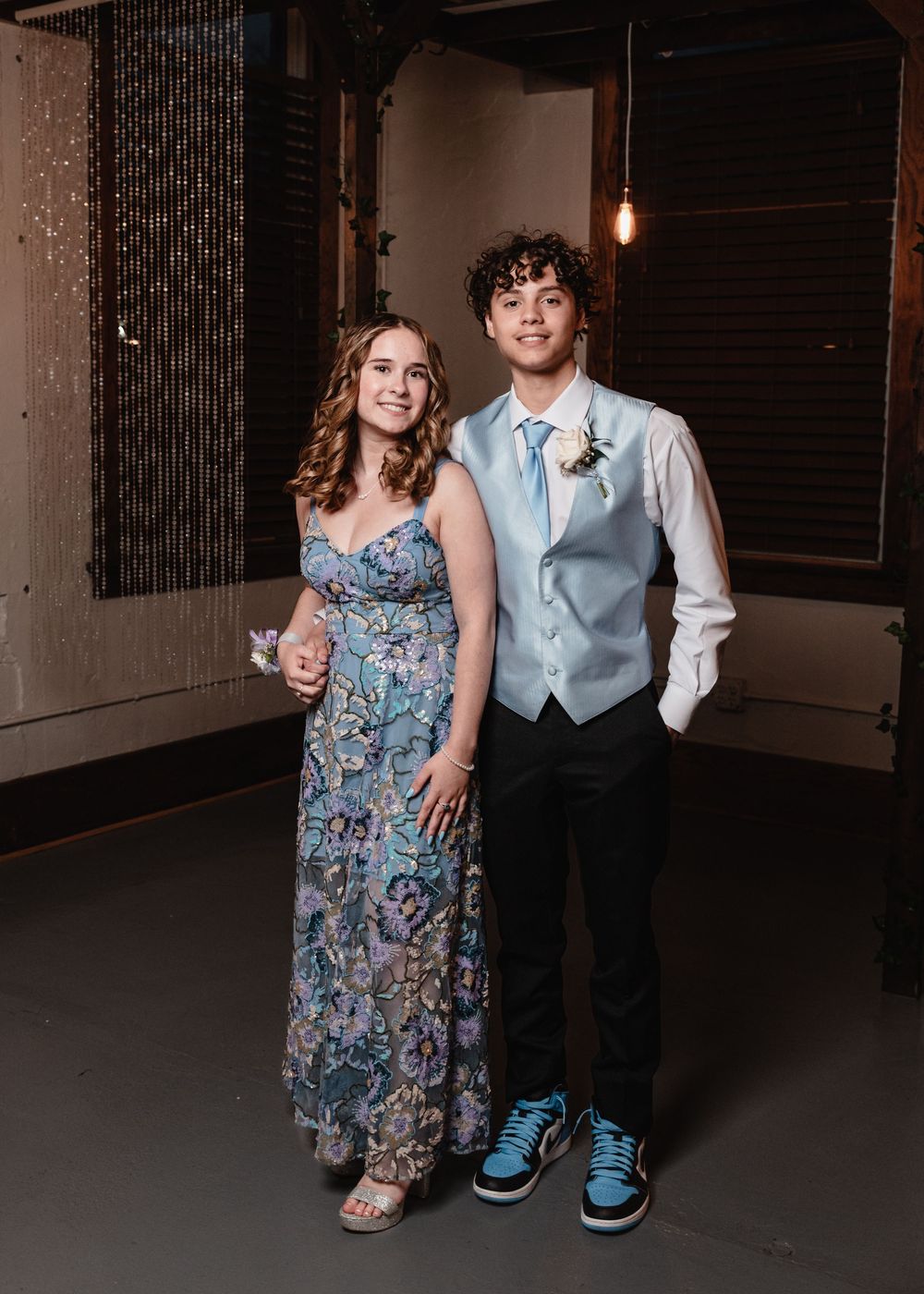 A couple poses together at a formal event wearing a blue floral dress and light blue vest with dress shoes.