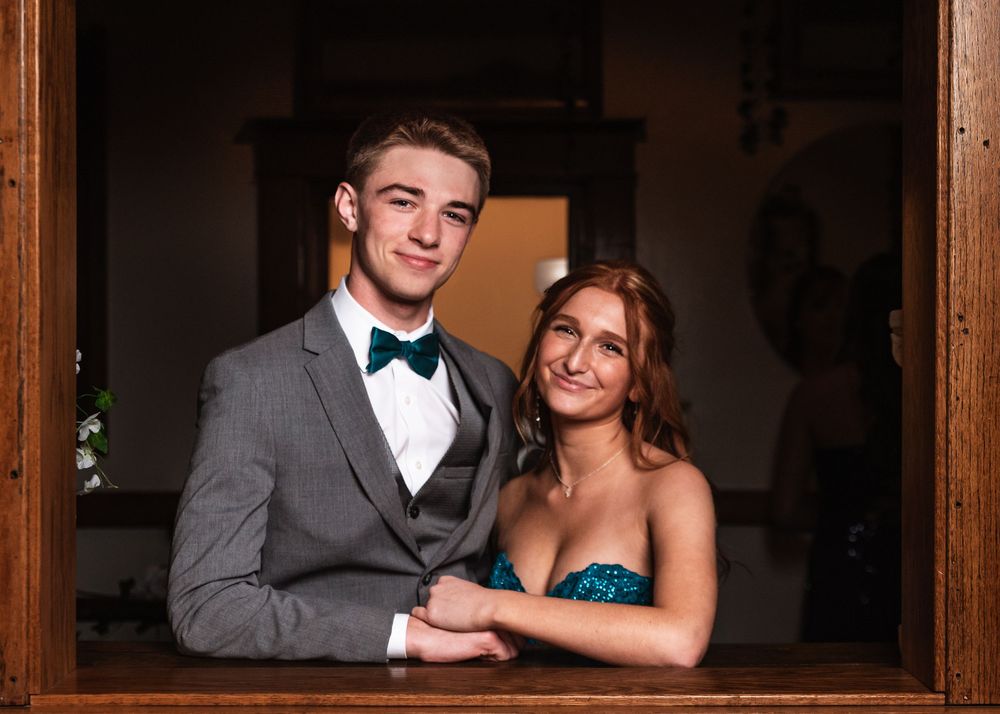 A smiling couple poses together for a formal portrait in elegant attire through a wooden window frame.