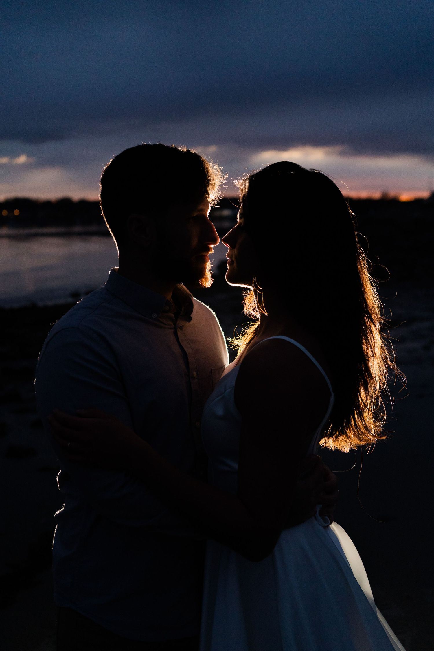 Silhouetted couple embraces at sunset by the water with dramatic backlit glow through their hair.