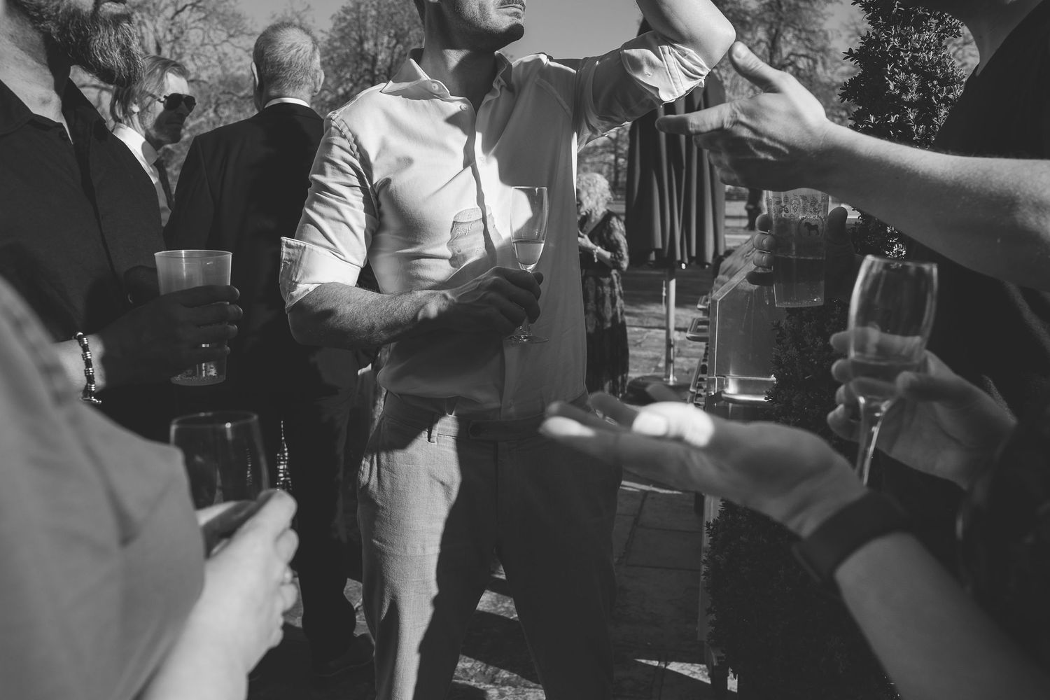 Black and white image of people holding drinks at an outdoor wedding reception with hands reaching across the frame.
