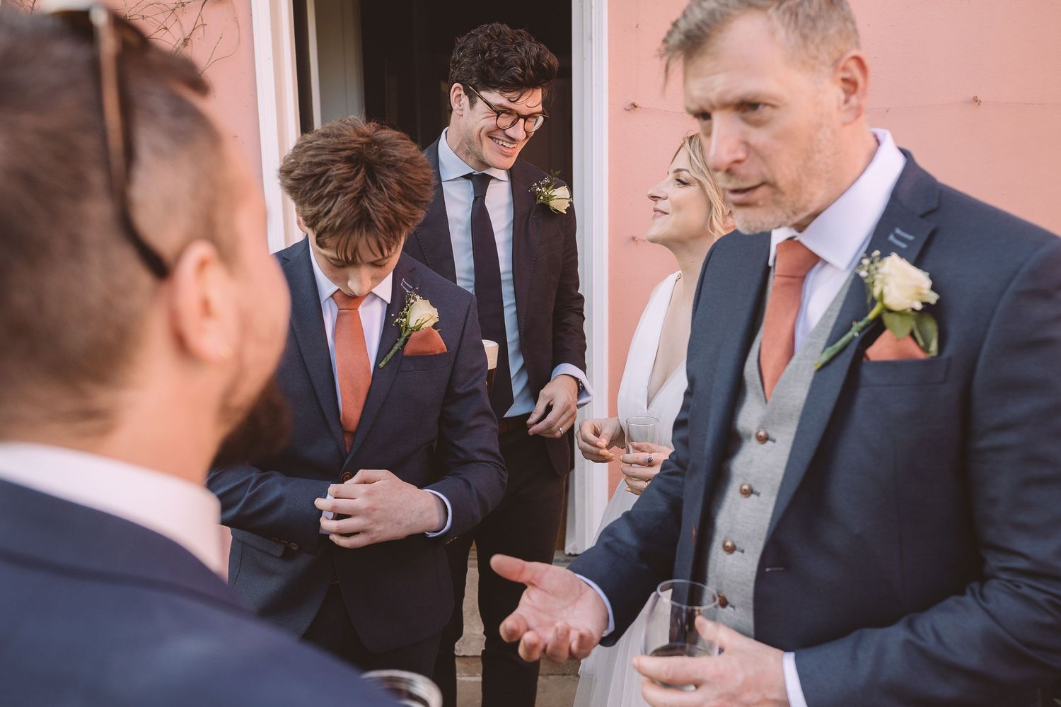 Groomsmen in navy suits with coral boutonnieres stand together against a pink wall during the wedding reception.
