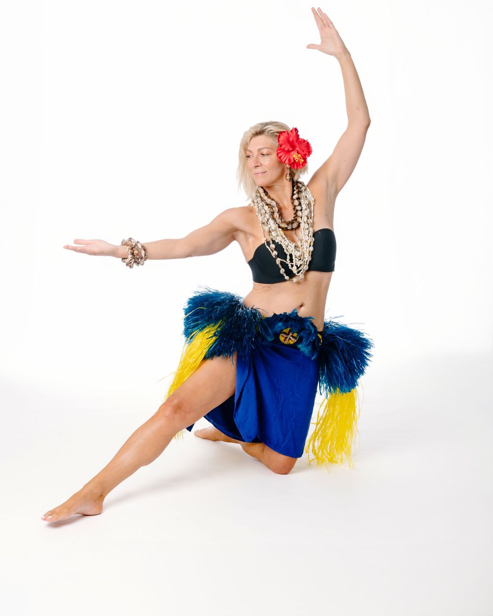Blonde hula dancer in blue feathered skirt and black top poses gracefully with red flower in hair.