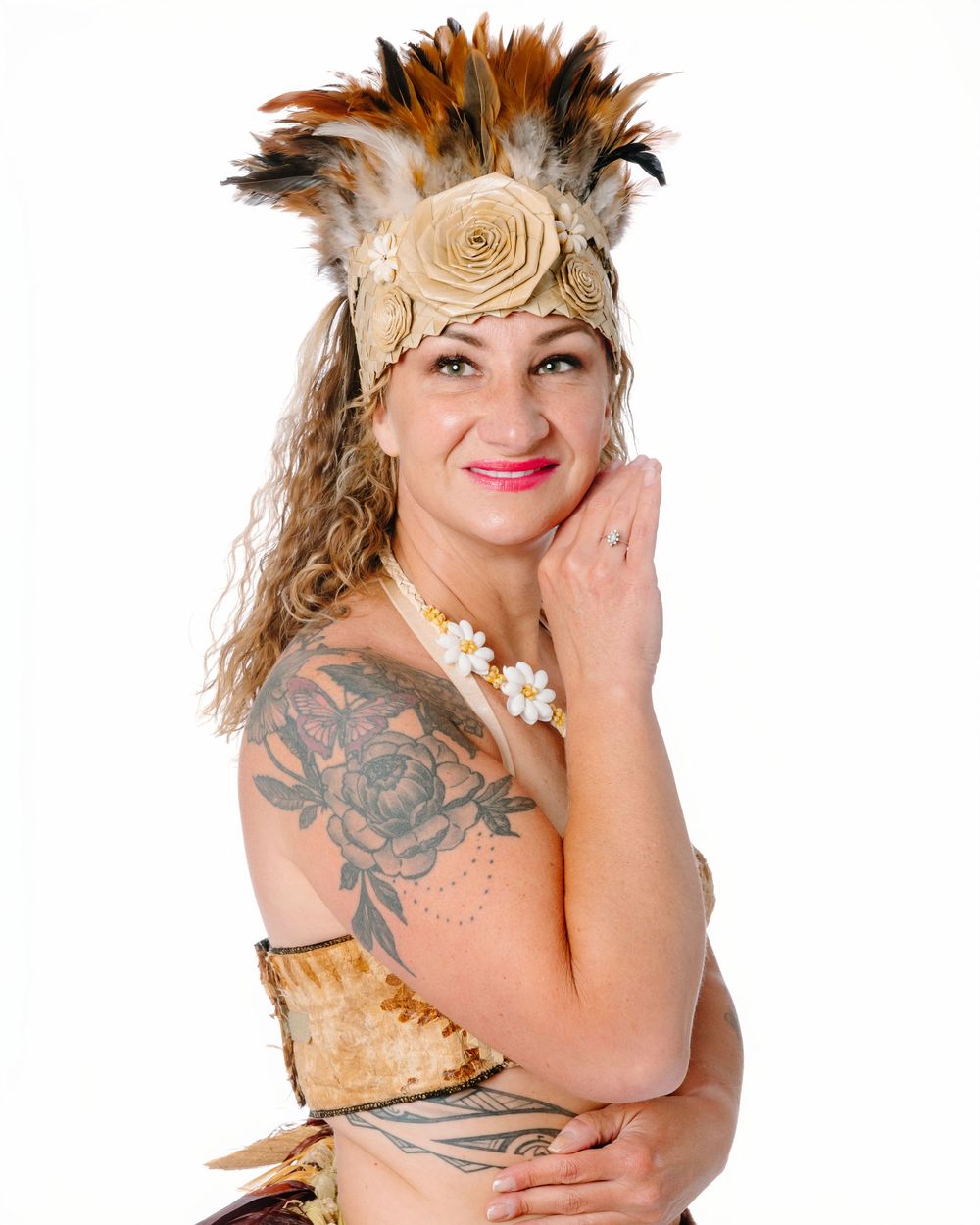 Woman in festive feather headdress with floral accents, tattoos, and beaded accessories poses confidently against white background.