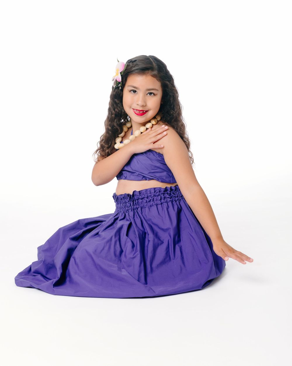 Young girl in purple crop top and skirt with pearl necklace and hair flower poses gracefully on white background.