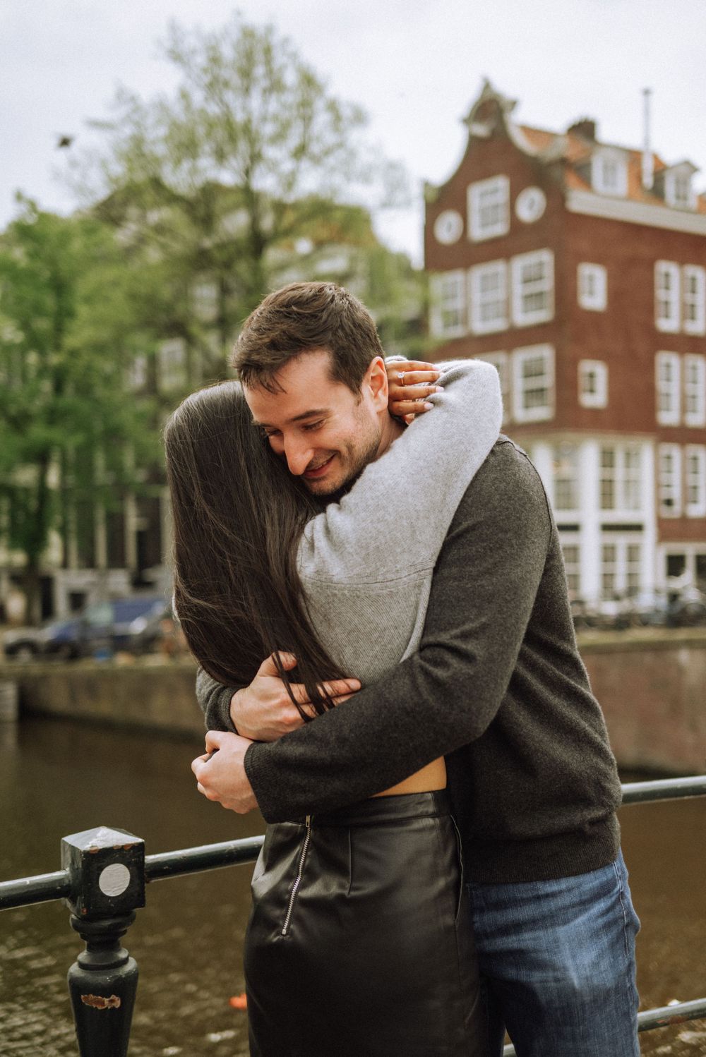 A joyful embrace along an Amsterdam canal with traditional Dutch architecture in the background.