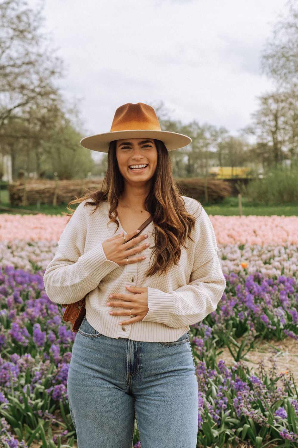 Person in brown hat and cream sweater posing joyfully in a field of purple flowers.