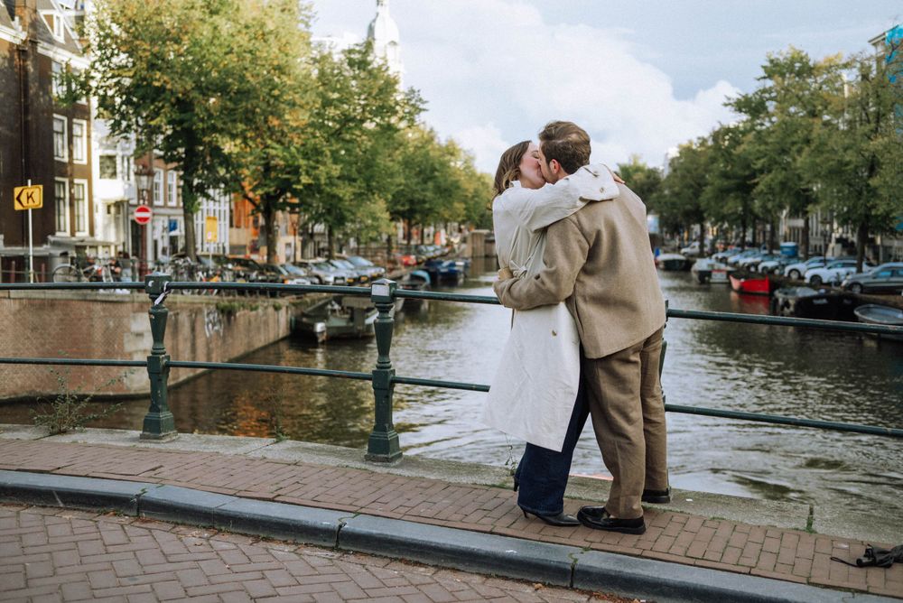 A romantic embrace by an Amsterdam canal with boats and historic buildings in the background.