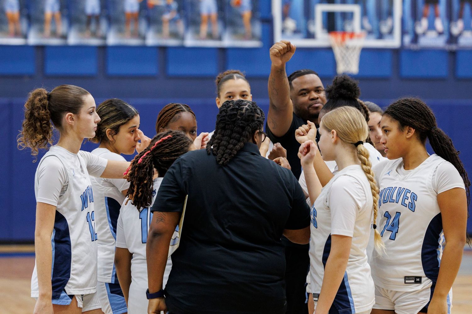 Basketball team huddles together during a timeout in a blue-themed gymnasium.