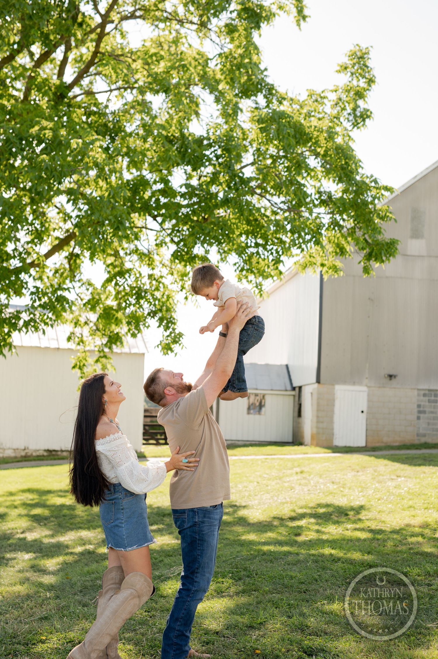 Parents lifting child in playful moment outdoors near white barn.