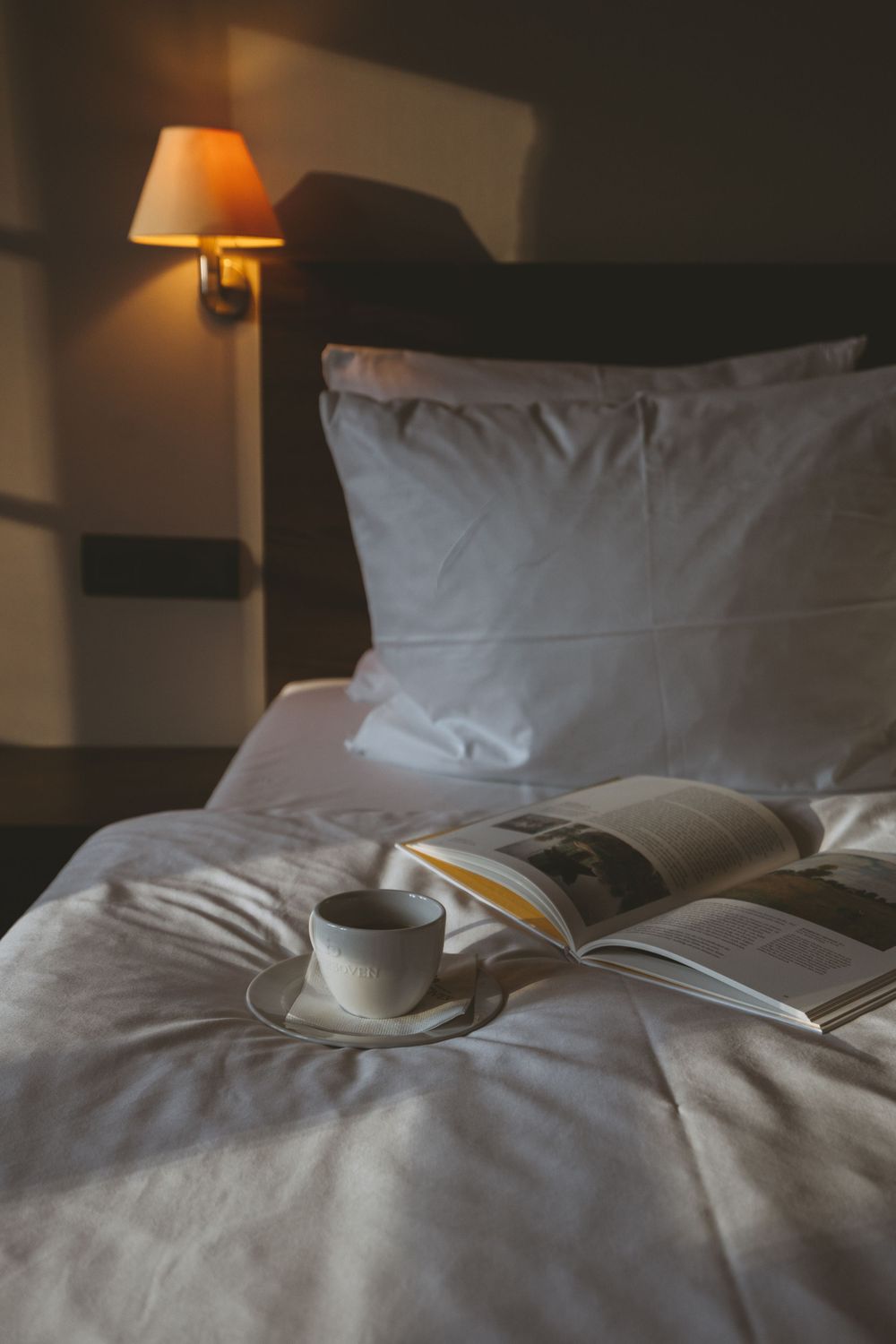 Open book and coffee cup on white bedding with warm lamp light creating a cozy reading atmosphere.