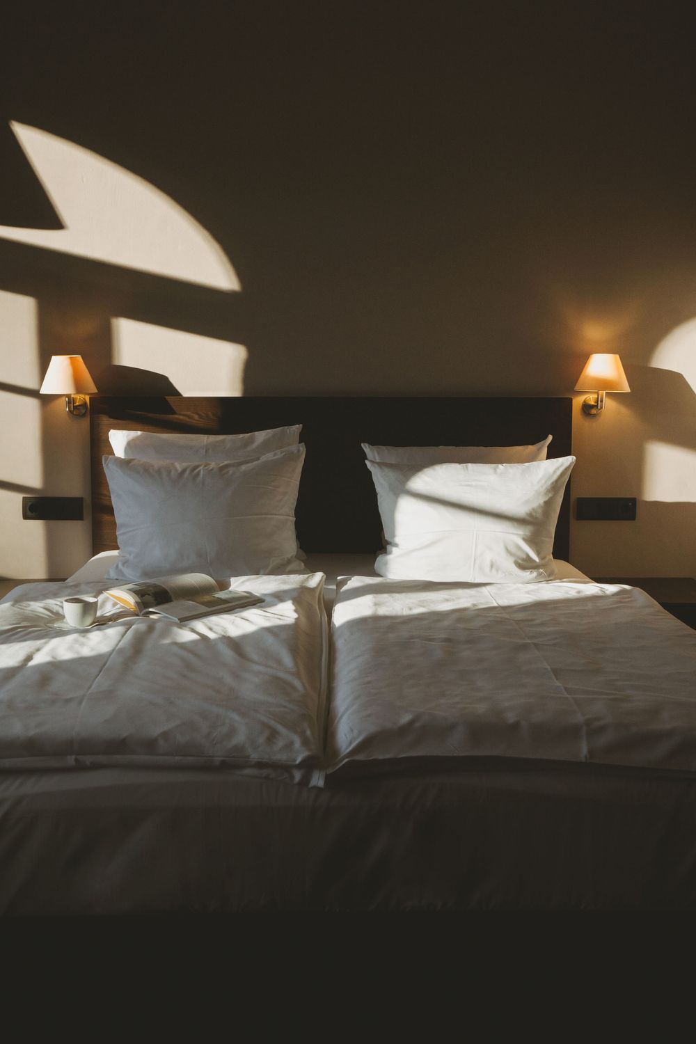 Modern hotel room interior with crisp white bedding and symmetrical bedside lamps casting geometric shadows.