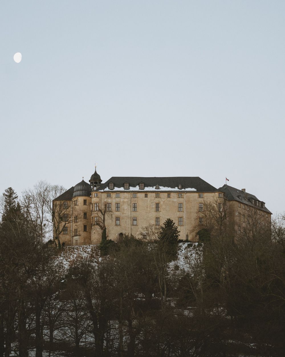 Castle-like stone building perched on hillside with moon visible in pale winter sky.