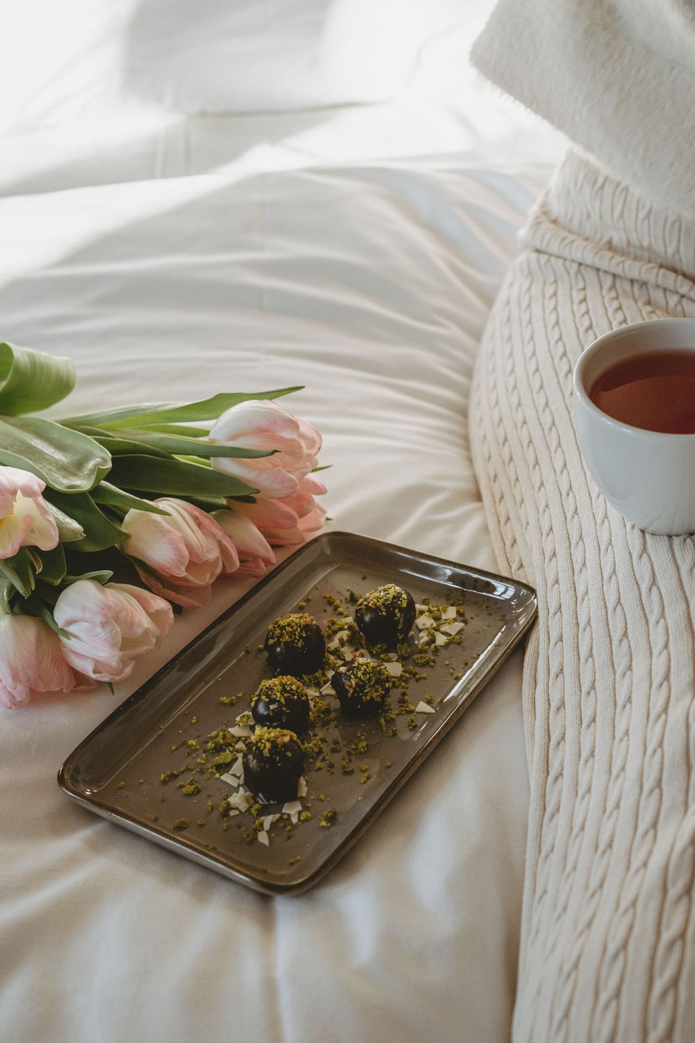 Pink tulips and a cup of tea arranged with dried herbs on white bedding.