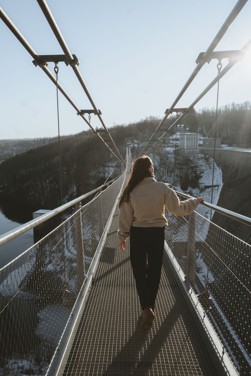 Person standing on suspension bridge overlooking snow-covered mountain valley.