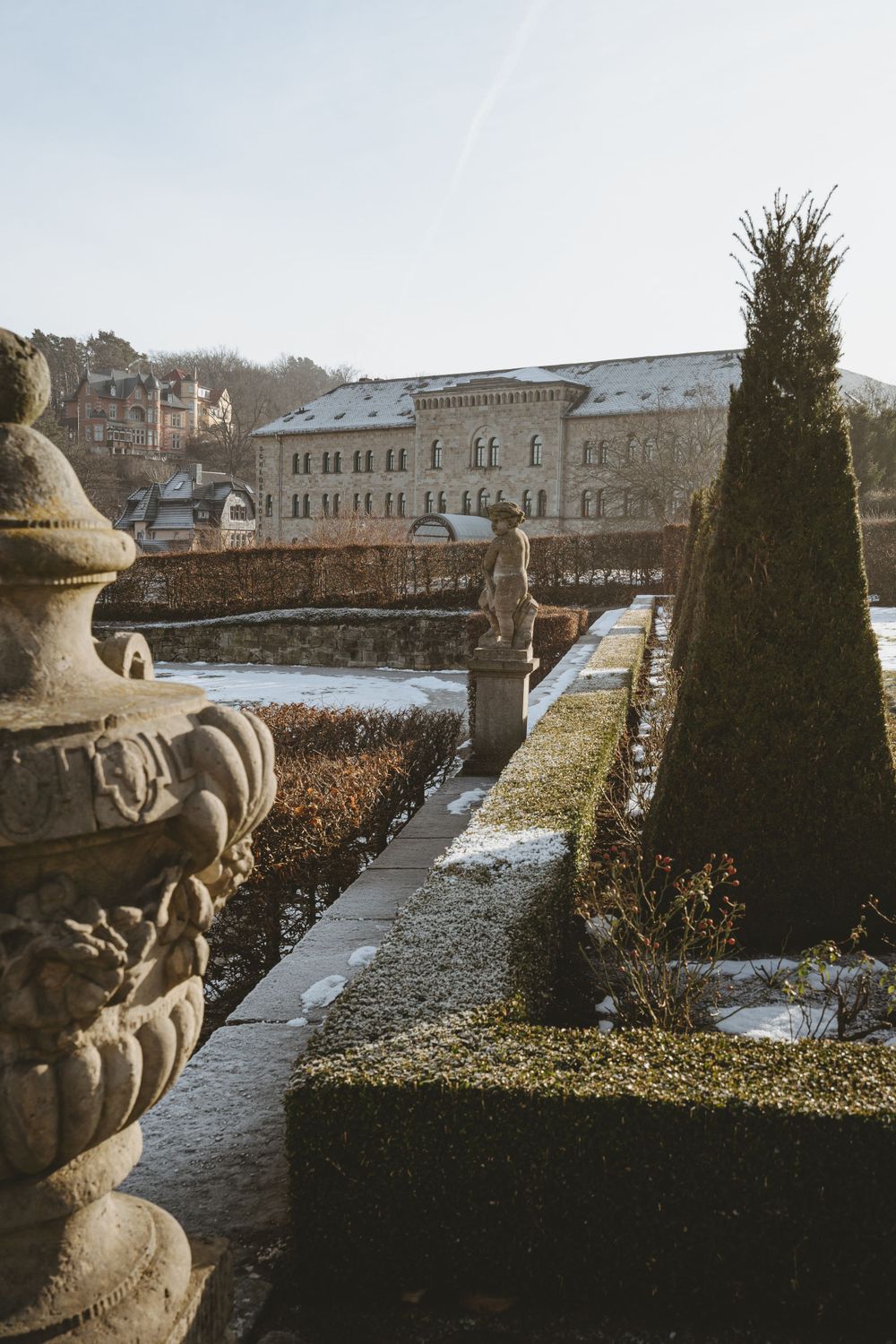 Snow-covered formal garden with ornate stone urns and geometric hedges leading to a historic building.
