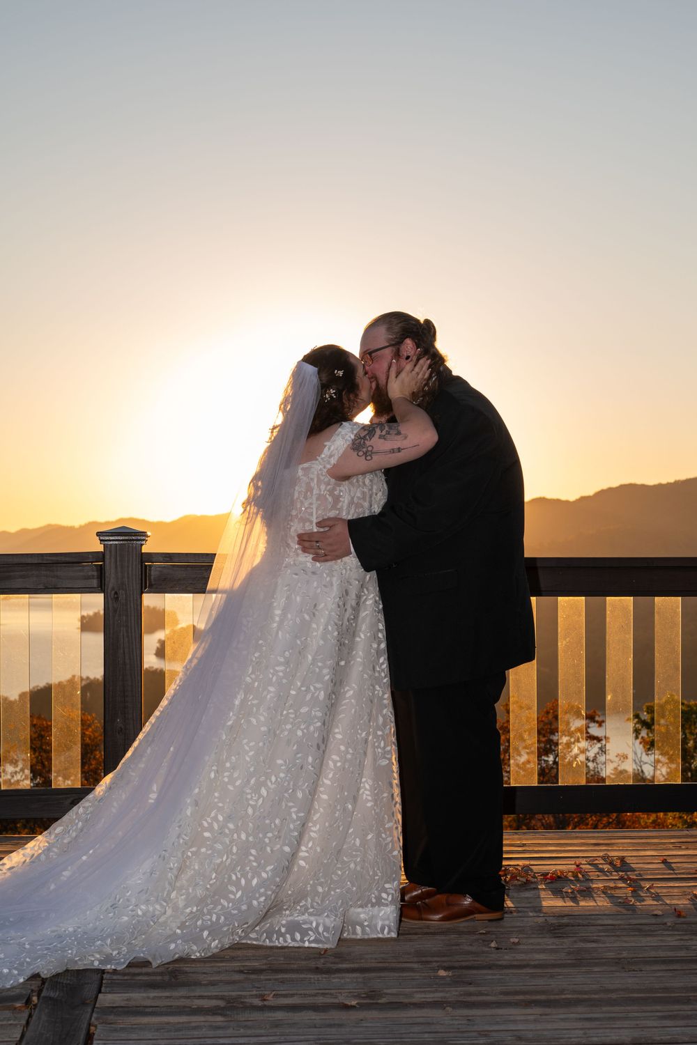 Sunset silhouette of newlyweds embracing on mountain deck with lake view.