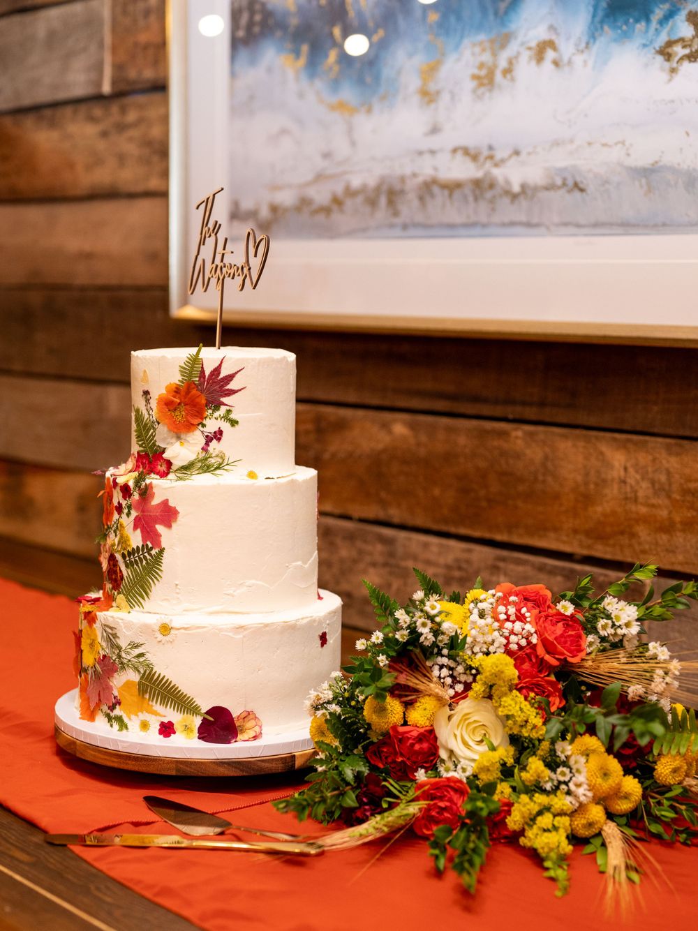 Three-tier white wedding cake decorated with autumn flowers and greenery beside a colorful bouquet.