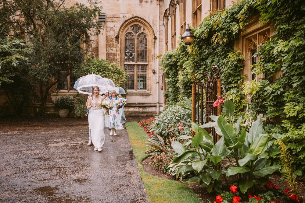Bridal group walking with umbrellas along gothic building pathway with ivy and flowers in rain towards chapel at Balliol College, Oxford.