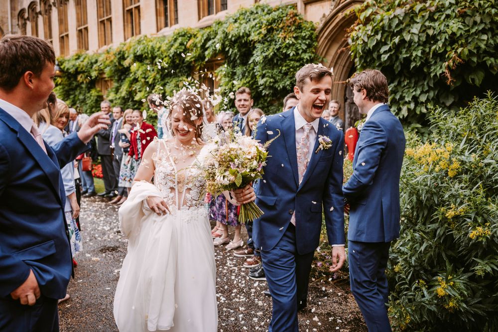 Newlyweds walk through shower of confetti in stone courtyard with wedding guests.