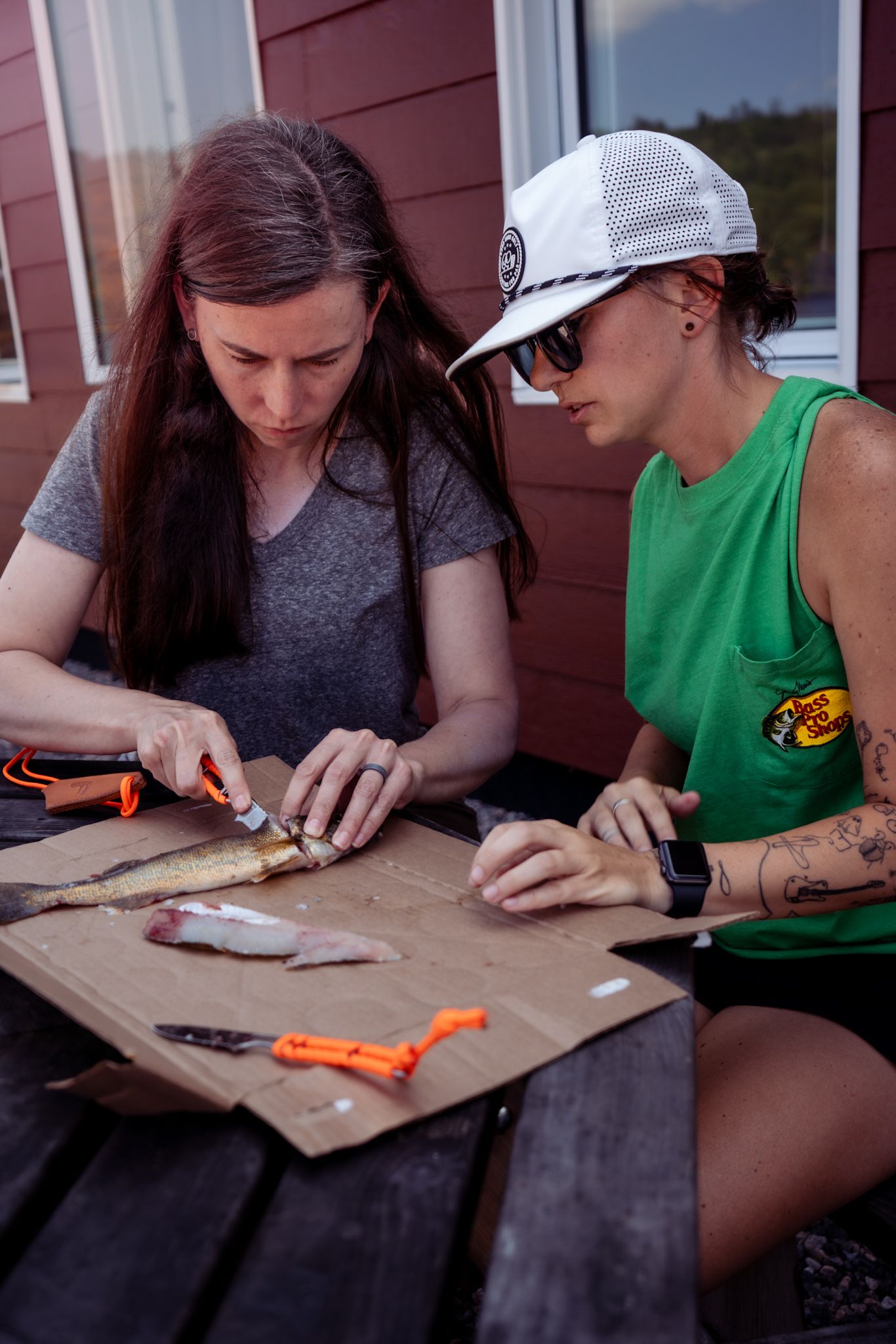 People sit at wooden table working with craft supplies near a red building on a sunny day.