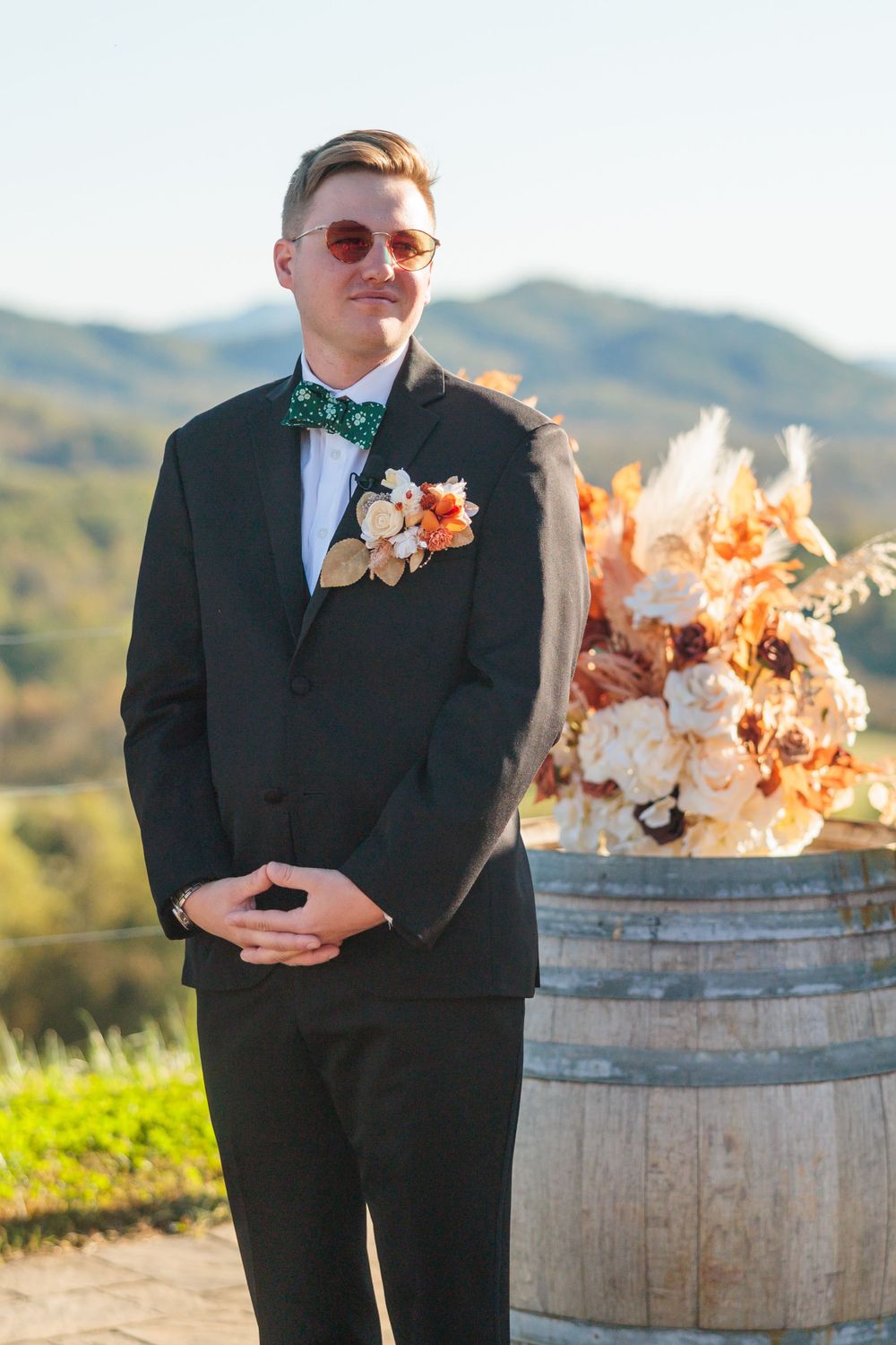 A stylishly dressed guest stands next to a rustic barrel with floral arrangements at an outdoor venue.
