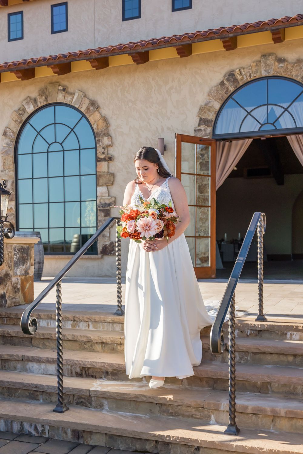 A bride in white holding a vibrant bouquet stands on stone steps of a Mediterranean-style building.