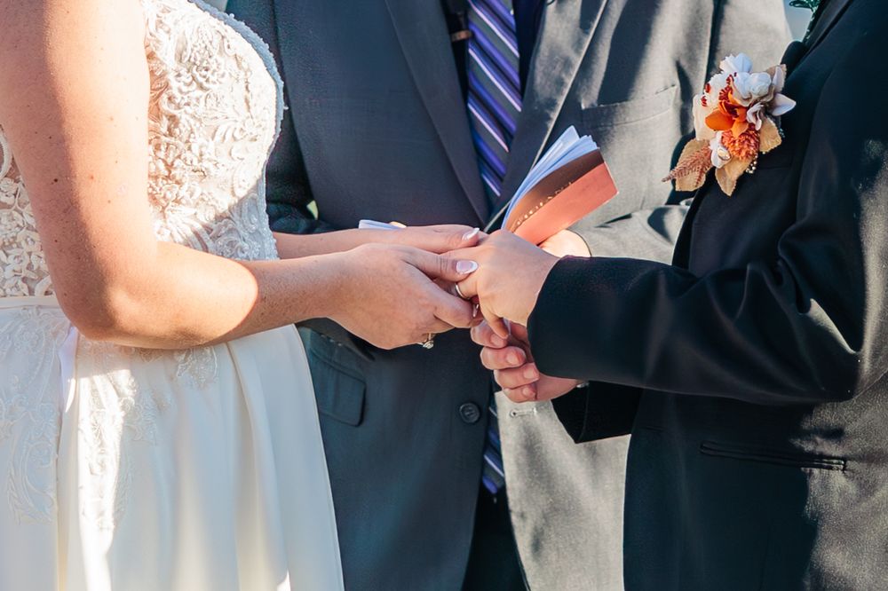 Close-up of hands exchanging wedding rings during ceremony with white lace dress and blue tie visible.