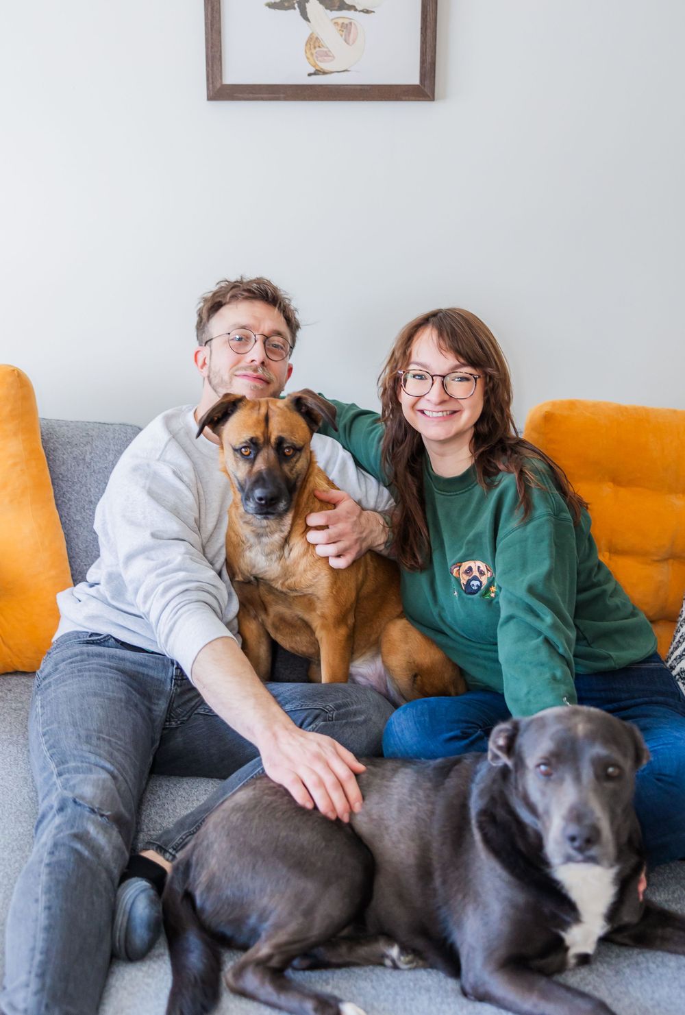 Family portrait with two dogs on grey couch under framed wall art.