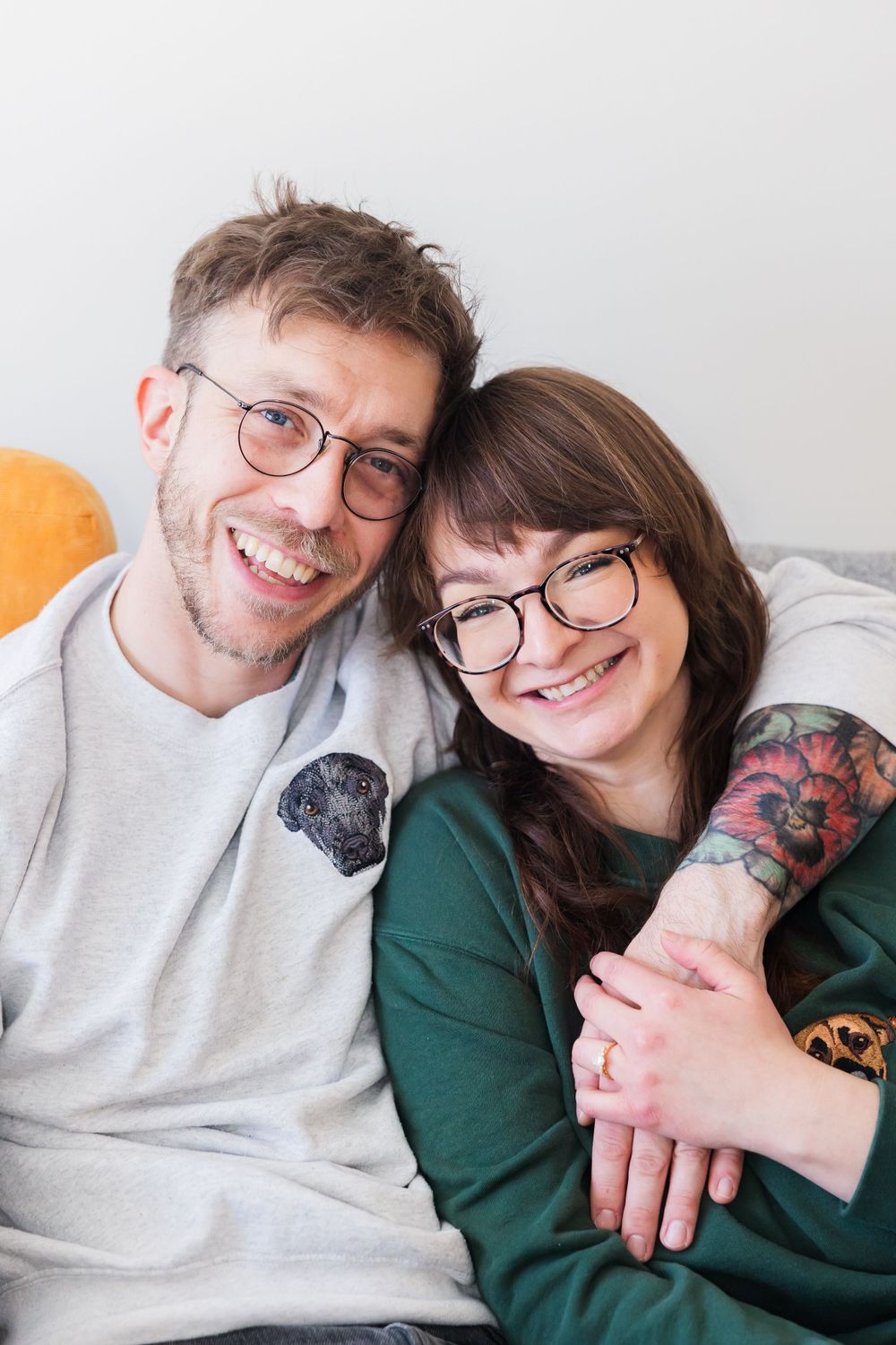 Couple wearing glasses smiles at camera while embracing on couch.