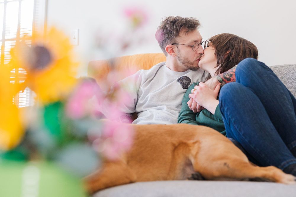 Couple shares intimate moment on couch with brown dog and spring flowers in foreground.