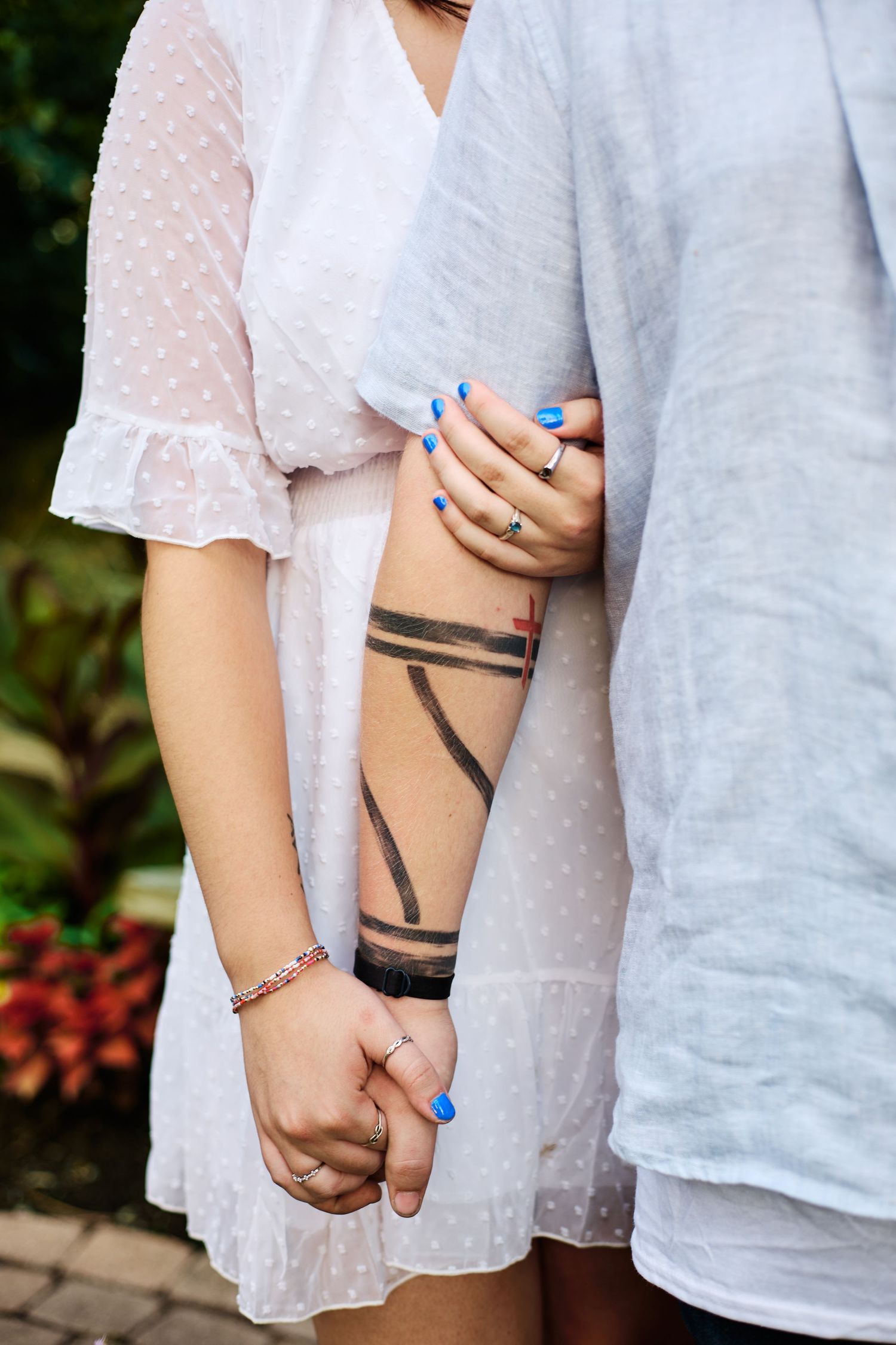 Close-up of hands with blue nails and geometric arm tattoo against white lace dress and gray clothing.